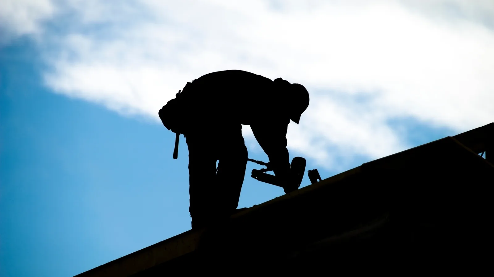 Backlit silhouette of a roofer using a nail gun on a sloped roof